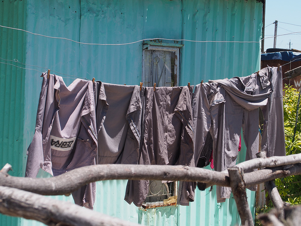 Work clothes hang on a clothesline outside a shack in an informal settlement that we photographed in Khayelitsha. Informal settlements are areas with makeshift dwellings crammed together. Residents do not have access to water in their homes or on their property. They must walk to get water from a communal water source and use communal toilets, which are often in disrepair, and the toilets unclean.