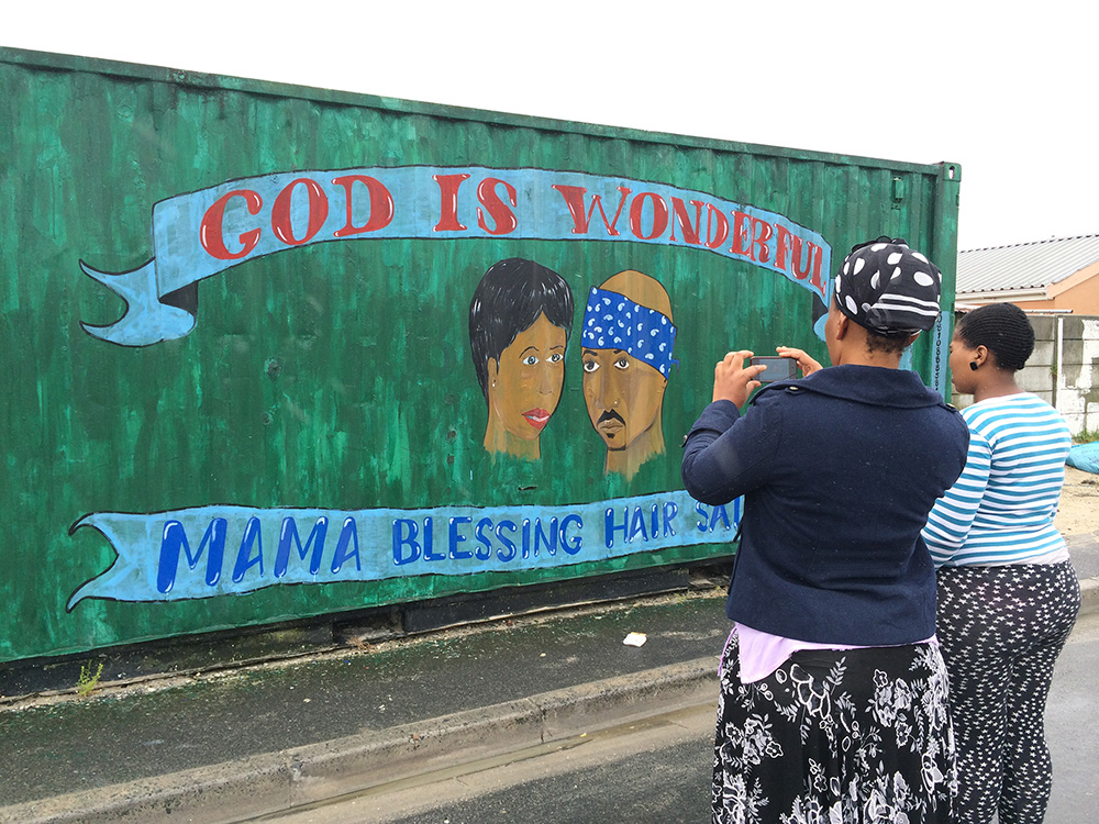 Yolanda Nkatula and Aviwe Dalingozi photograph the mural on a shipping container, home to Mama Blessing Hair Salon in Khayelitsha. The Townships have become like cities and are made up of small commercial shopping malls and independently owned businesses. These businesses sell their goods and services in shacks, shipping containers or simply on the sidewalk. With high unemployment rates in Khayelitsha, it is necessary to find economic opportunities. Most of the containers and shacks are painted with colourful murals with many honouring their strong beliefs.