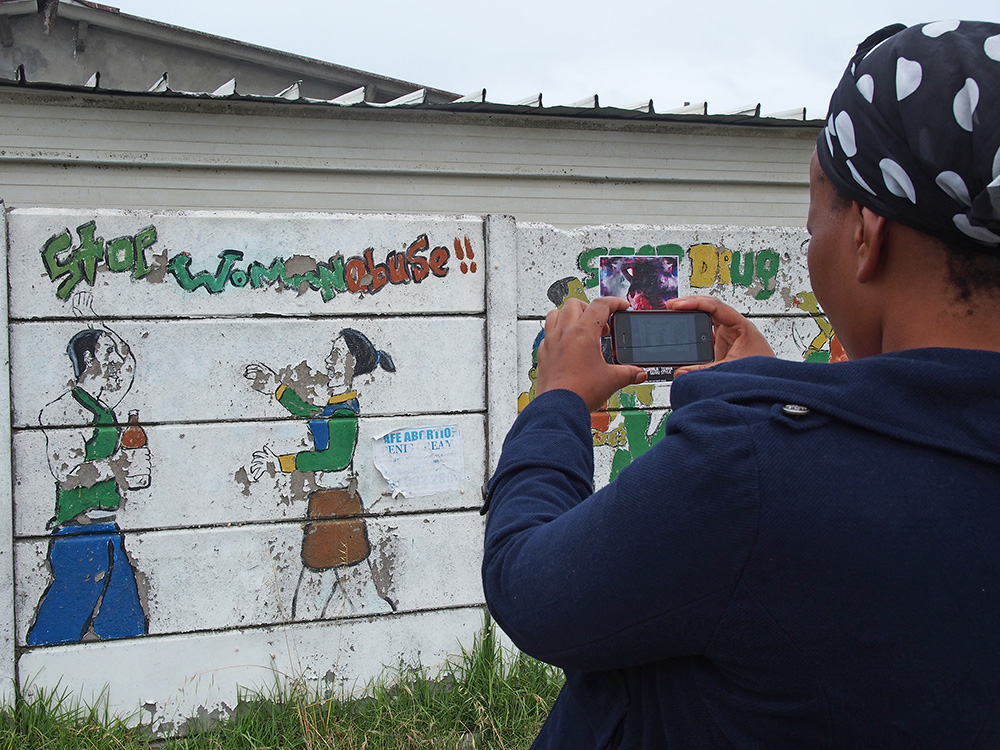 Yolanda Nkatula, a married mother of 2 pauses to photograph a ‘Stop Woman Abuse’ mural on Walter Sisulu Road in Khayelitsha in November 2013. Many of the fences are painted with street art and showcase anything from advertising a local business to addressing social issues.